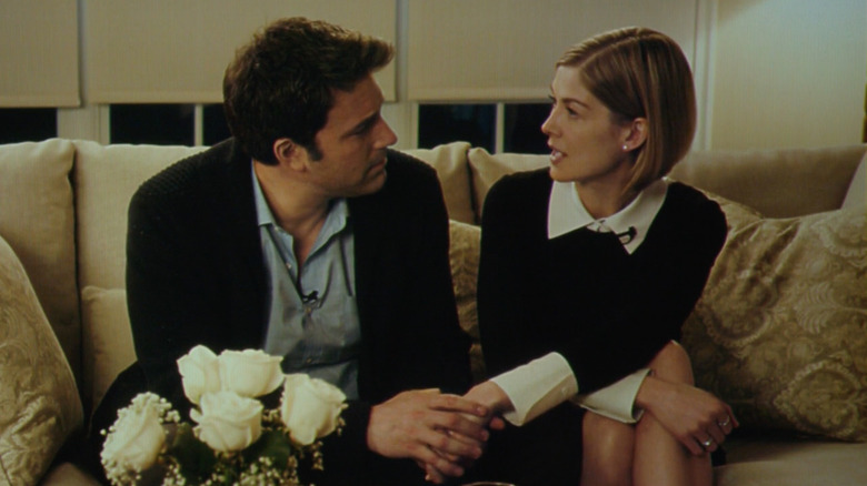 Rosamund Pike as Amy Dunne in Gone Girl, wearing a black top with a white collar and cuffs, next to Ben Affleck as Nick Dunne