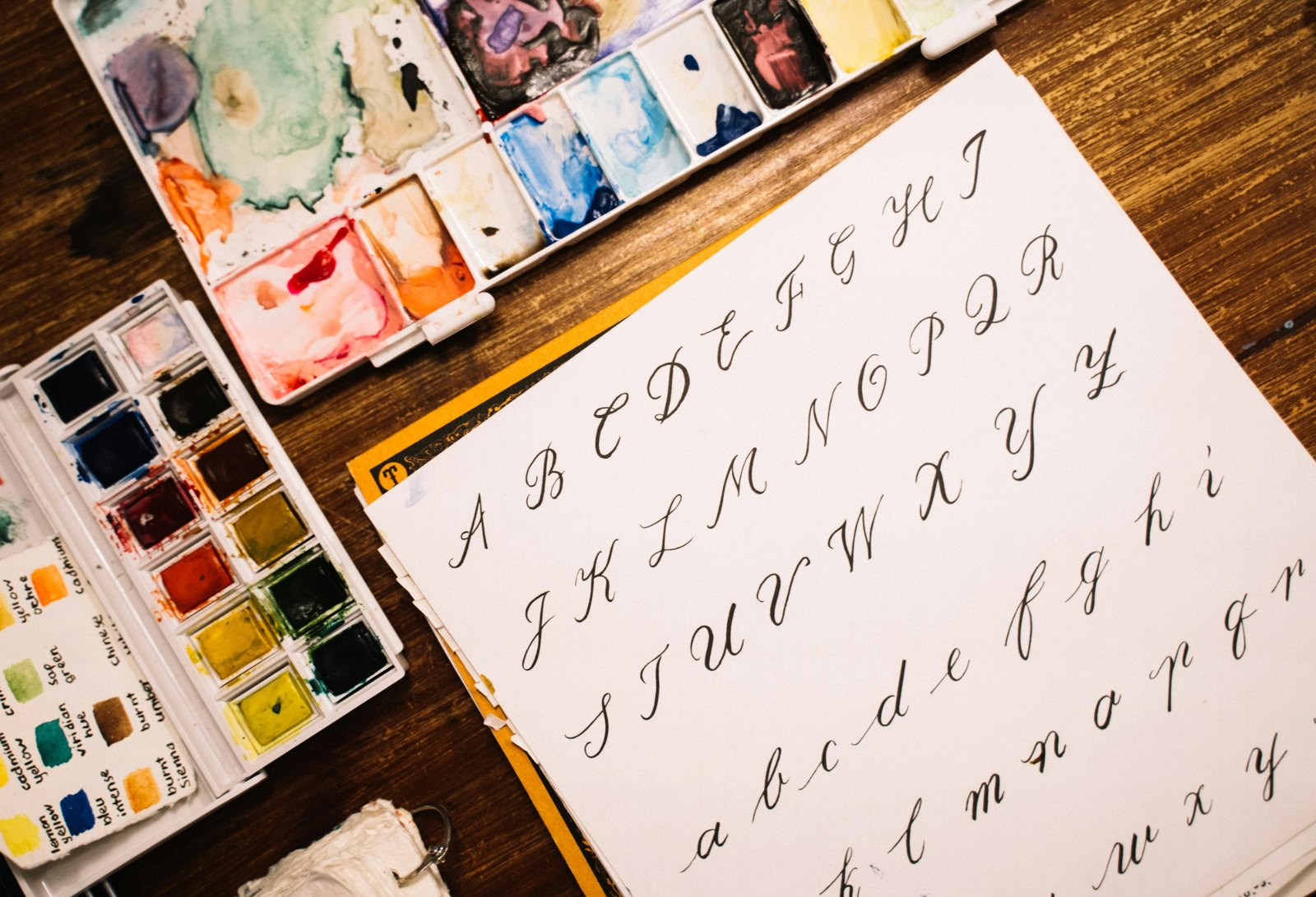 Brush lettering alphabet practice sheet next to watercolor palettes, a hand lettering workshop gift idea